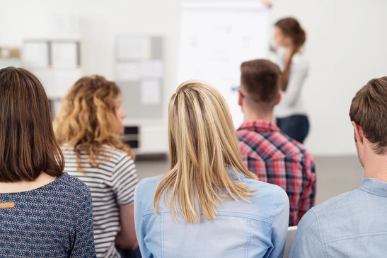 Salle de formation moderne avec groupe d'adultes assis face à une formatrice debout devant tableau blanc