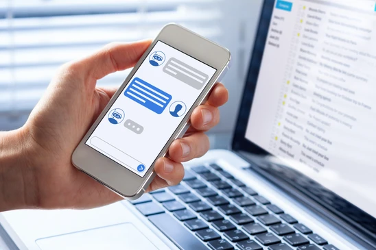 Main tenant un smartphone affichant une interface de chatbot conversationnel avec bulles de messages bleus et gris, icônes de profils utilisateurs