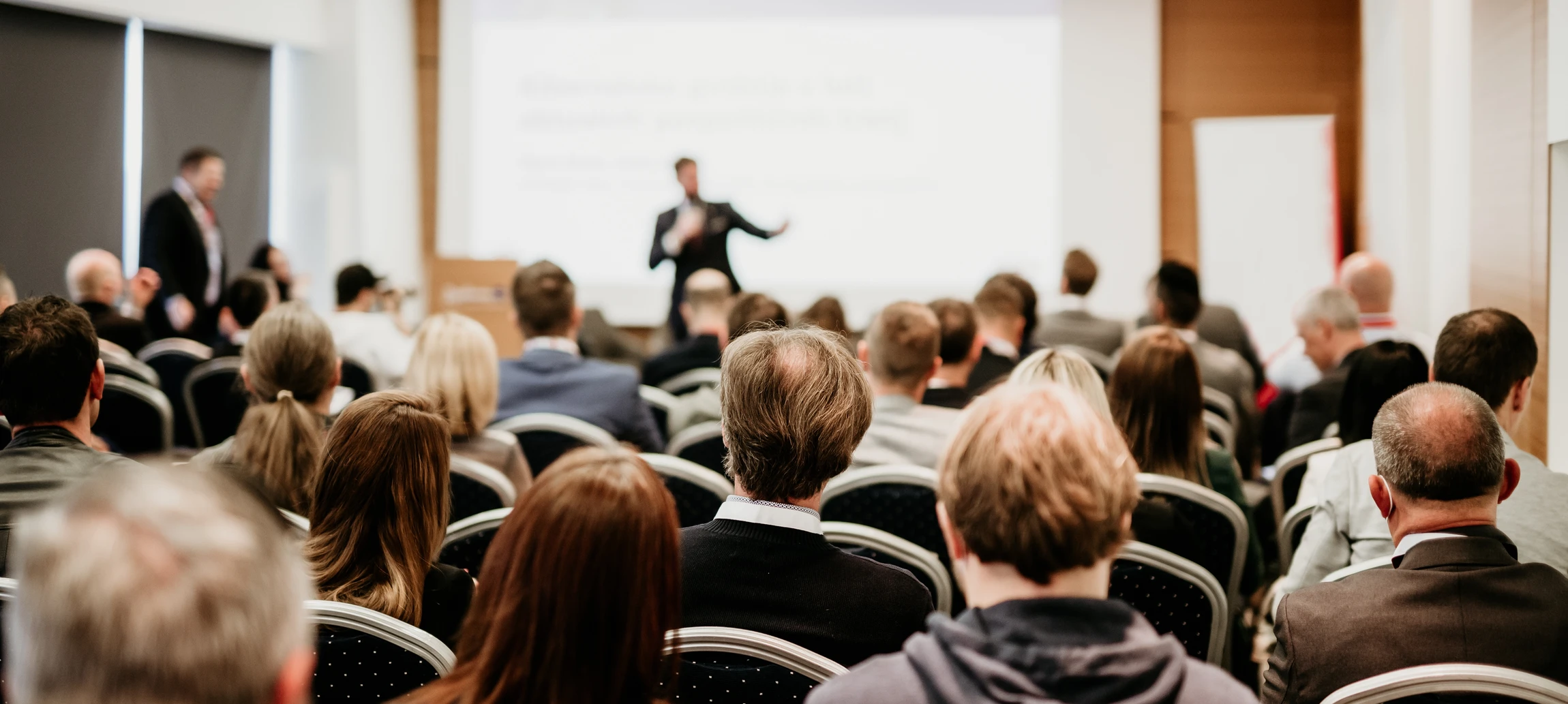 Photographe professionnelle montrant un formateur en train de faire une présentation devant un petit groupe de professionnels dans une salle de formation moderne et lumineuse. Le formateur, en tenue professionnelle, interagit avec son audience en pointant vers un écran affichant du contenu relatif à l'intelligence artificielle. L'ambiance doit être professionnelle, engageante et authentique, mettant l'accent sur la transmission de savoir et la collaboration. Lumière naturelle, environnement moderne et épuré.